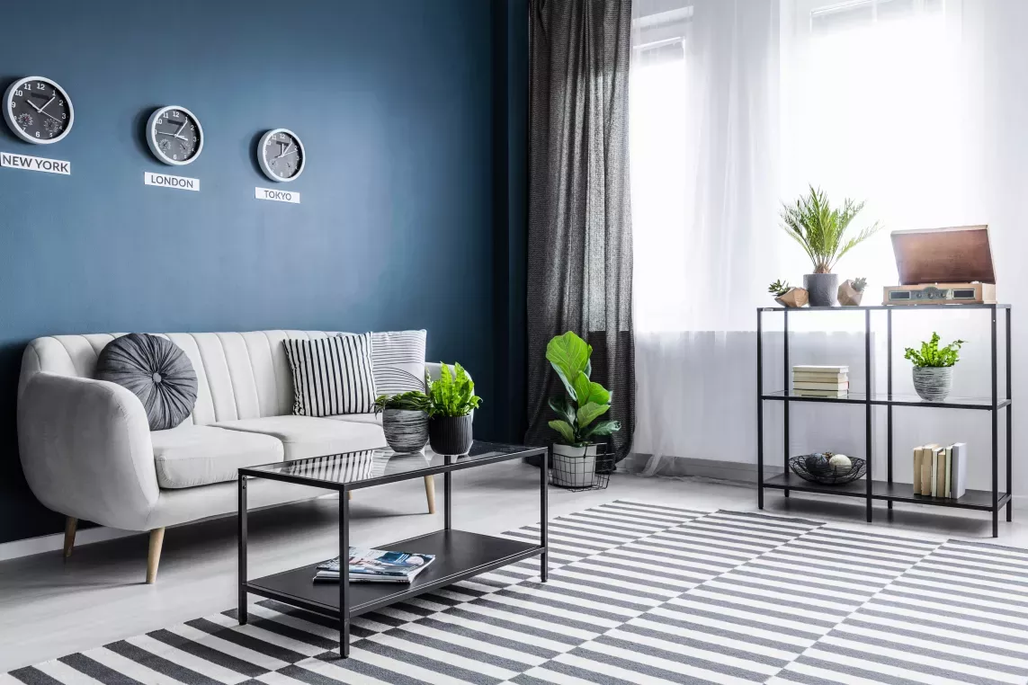 A modern living room featuring a white sofa with striped and circular pillows, a glass coffee table with plants, a striped rug, and a shelving unit adorned with books, plants, and a vintage record player, all set against a deep blue accent wall with three world clocks.