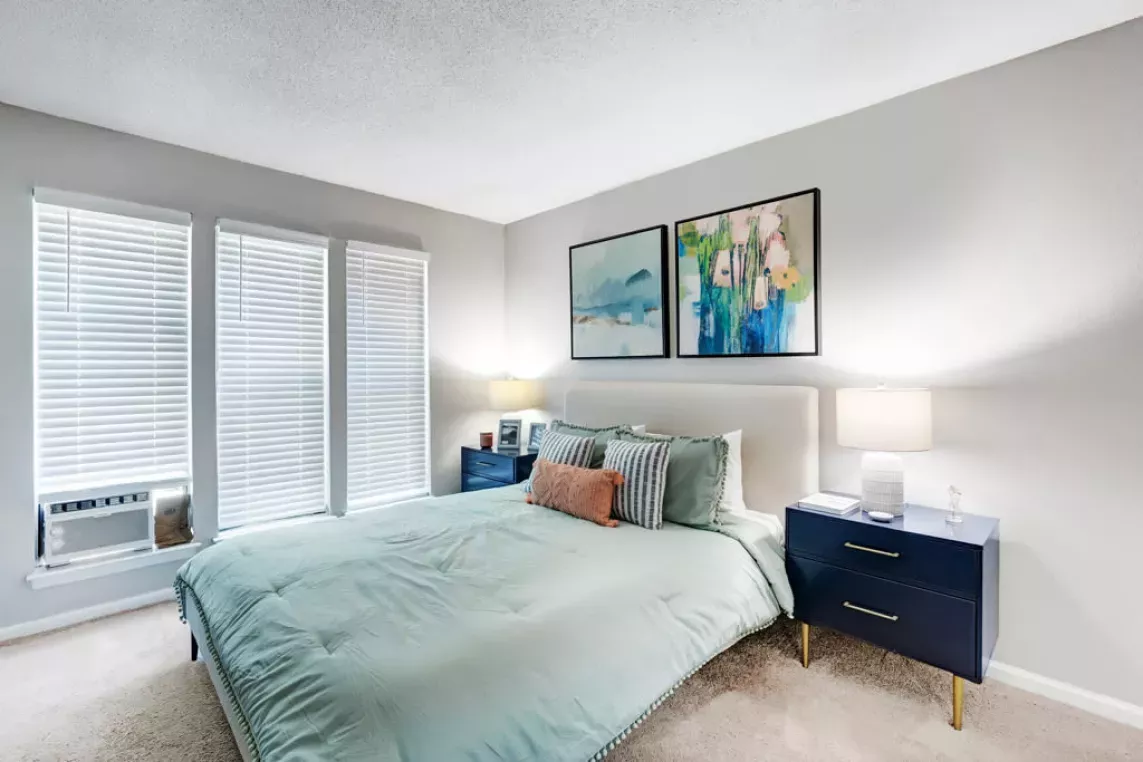 A bright bedroom featuring a neatly made bed with pastel bedding, modern navy nightstands, and abstract wall art above.