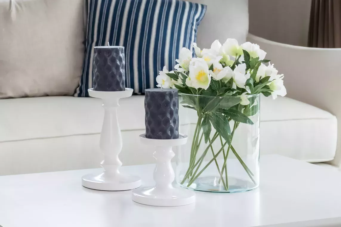 A cozy living room with a white coffee table featuring two dark candles on white holders and a vase of fresh white flowers, set against a soft beige couch with striped pillows.