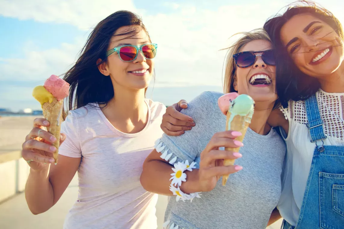 Three friends enjoying ice cream cones and laughing together at the beach.