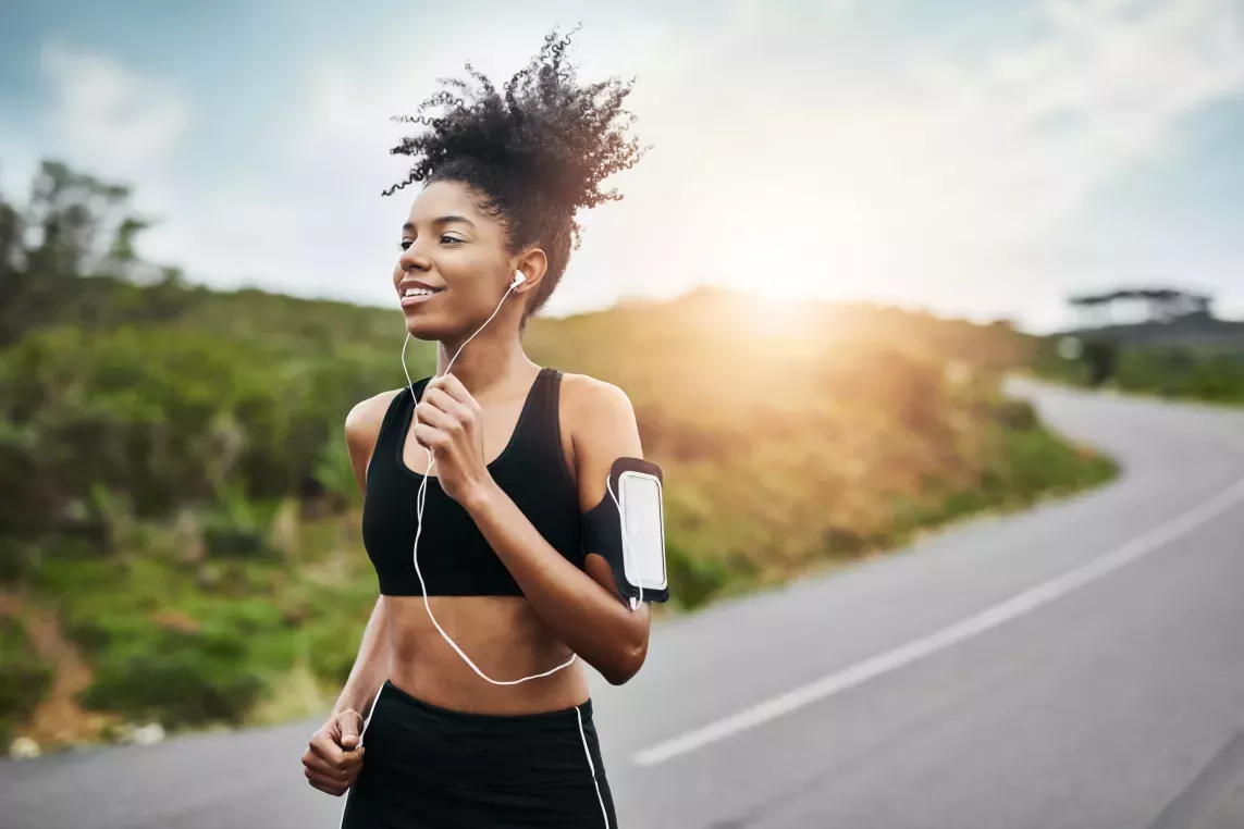 A woman jogging along a winding path while listening to music with the sun shining brightly in the background.