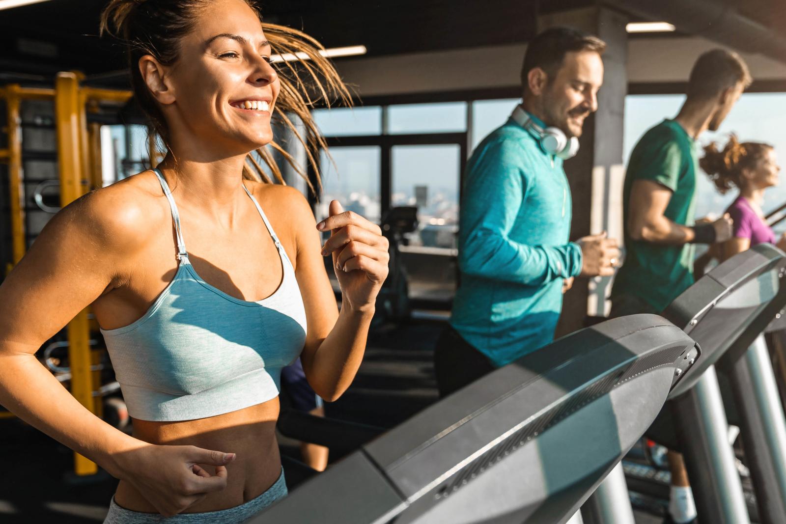 A group of individuals working out on treadmills in a brightly lit gym, featuring large windows and a variety of fitness equipment in the background.