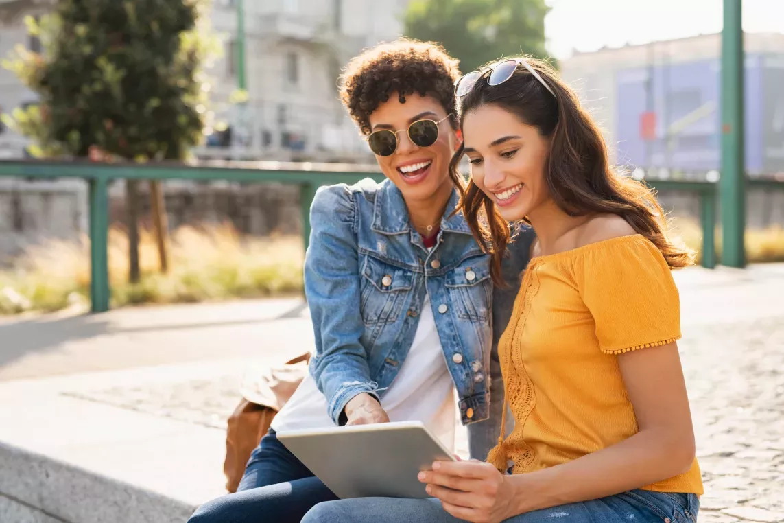 Two women are happily sitting outdoors, sharing a moment while looking at a tablet together on a sunny day.