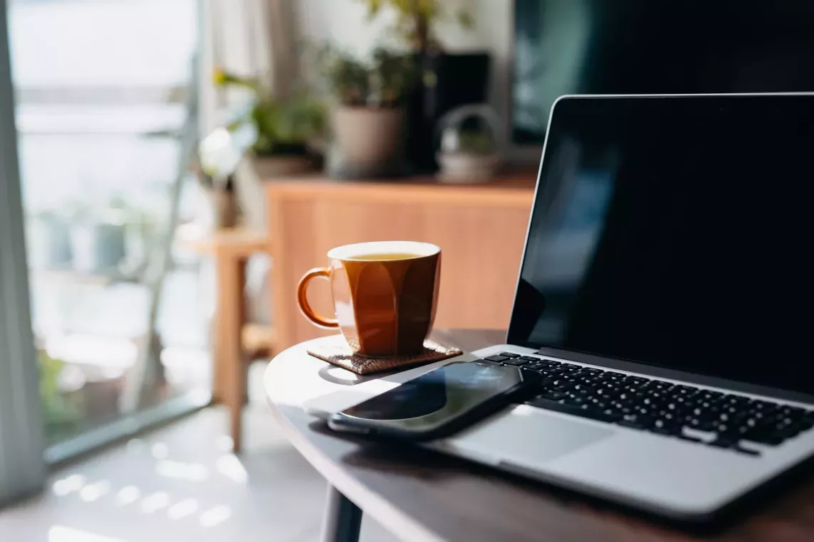 A cozy home workspace with a laptop, smartphone, and a cup of tea on a wooden table, surrounded by natural light and indoor plants in the background.