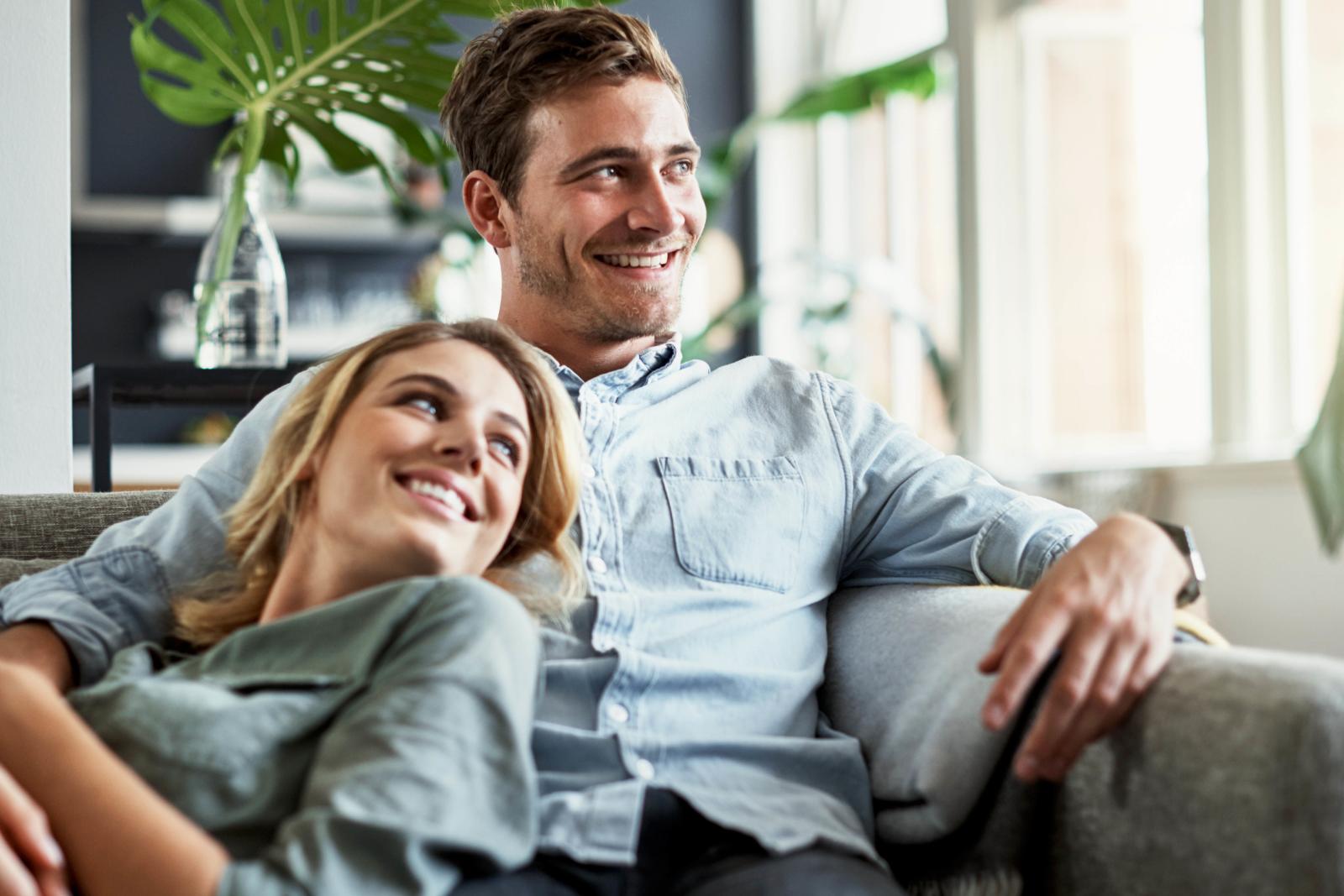 A smiling couple relaxes together on a cozy sofa in a bright, modern living space with greenery and natural light.