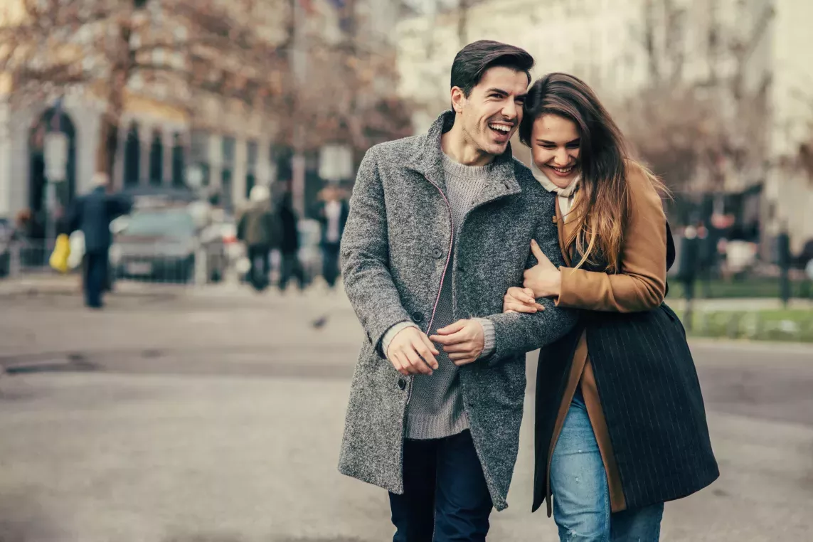 A cheerful couple walking arm in arm through a city park on a crisp day, surrounded by blurred urban scenery and trees.