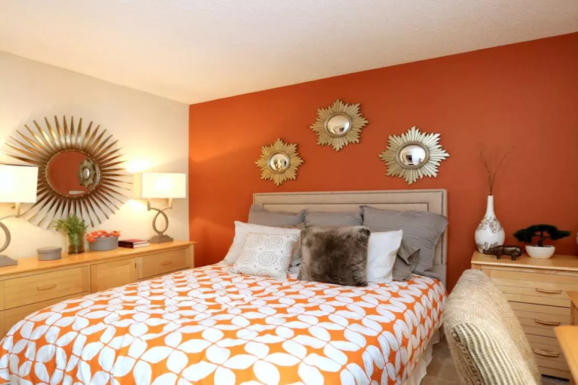 A stylish bedroom at Norcross Crossings Apartments features a bold orange accent wall, elegant decor with sunburst mirrors, and cozy furnishings for a warm and inviting atmosphere.