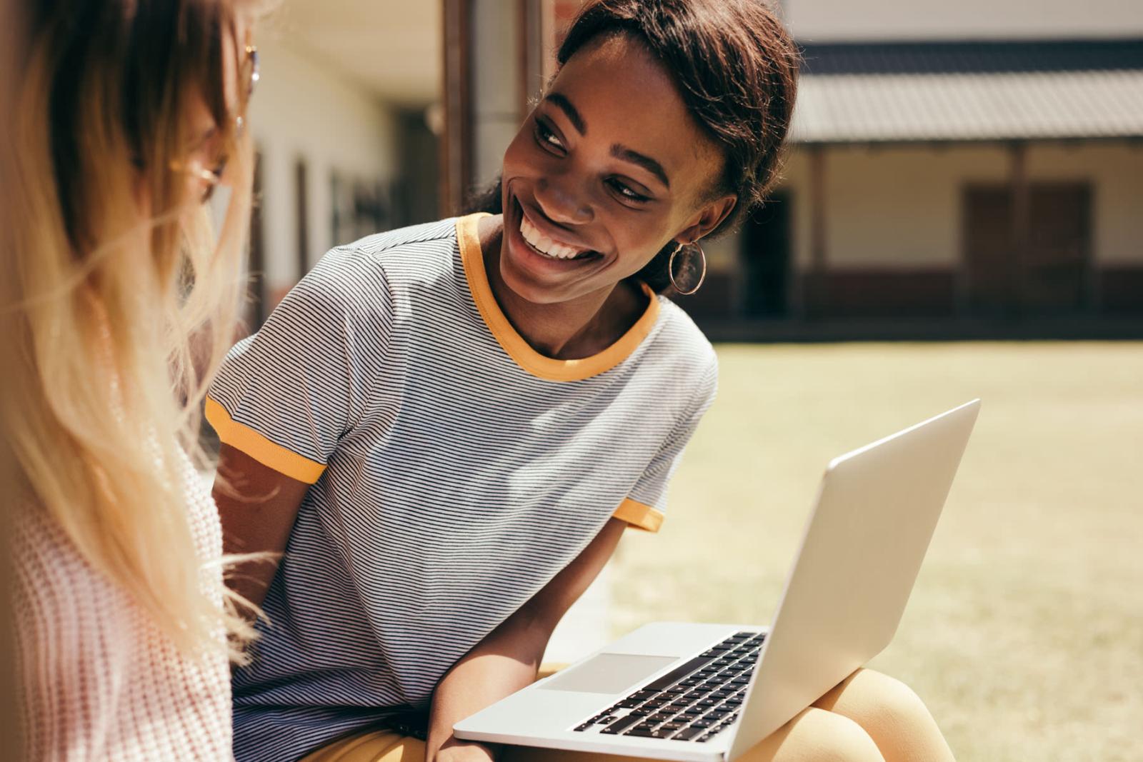 Two young women sitting outdoors, one with a laptop, smiling and engaging in conversation, highlighting the apartment community's proximity to educational opportunities and collaborative spaces.