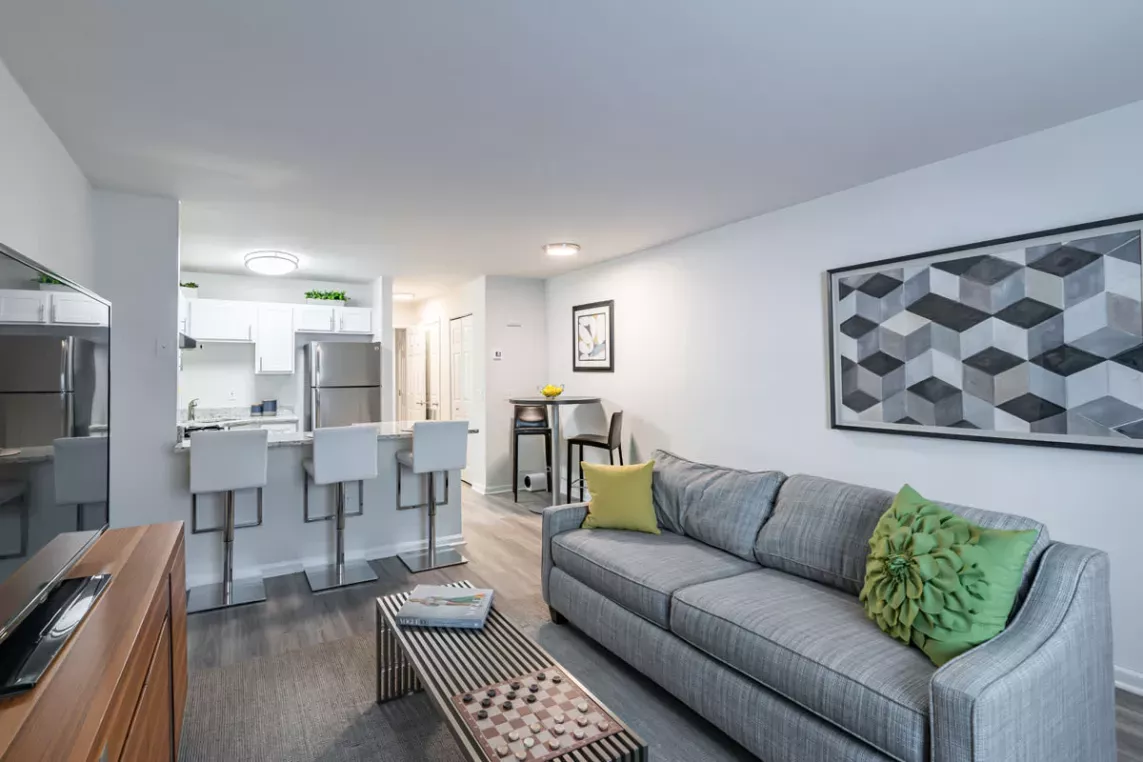 A modern living space featuring a cozy gray couch with green accent pillows, a geometric wall art piece, and a kitchen area with white cabinets and stainless steel appliances in the background.