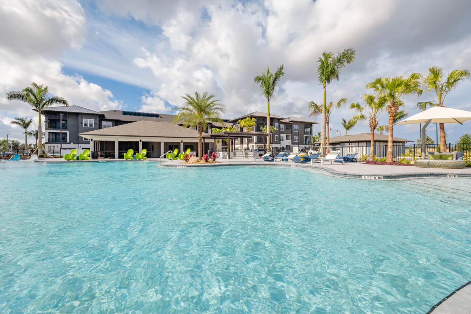 A spacious resort-style pool surrounded by lush landscaping, lounge chairs, and palm trees under a bright blue sky.