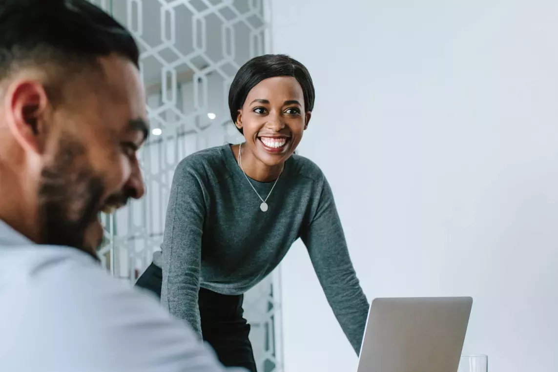 Two colleagues are engaged in a meeting, with the woman smiling as she stands near a laptop while the man is seated, smiling in the background.