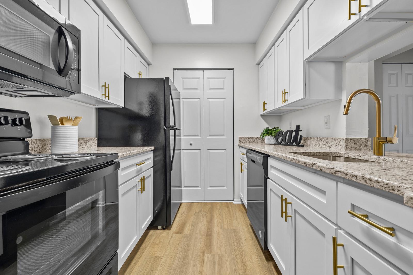 A modern galley-style kitchen featuring white cabinetry with gold handles, granite countertops, black appliances, and a stylish gold faucet, illuminated by a bright overhead light.