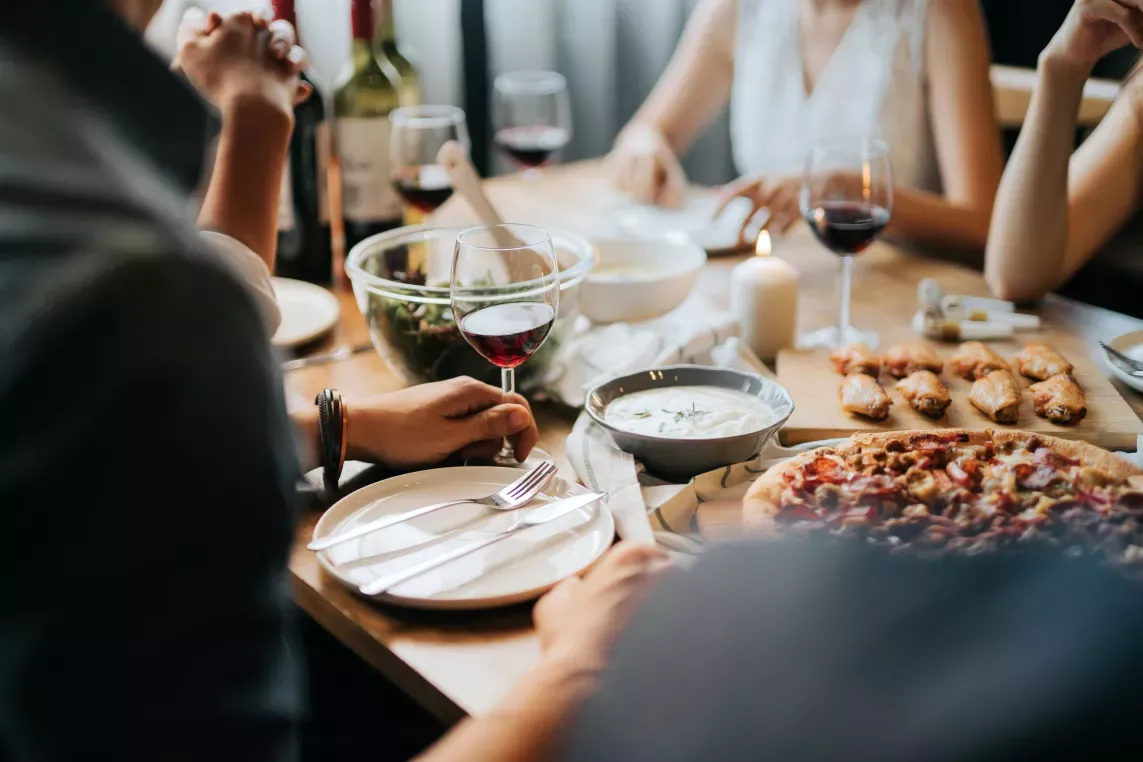 A cozy dinner gathering with friends, featuring red wine, pizza, chicken wings, and fresh salad on a warmly lit dining table.