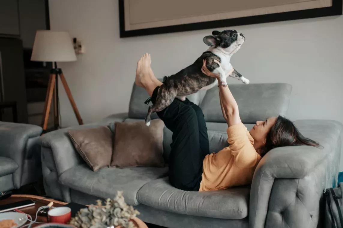 A woman lays on the couch while holding her dog in the air like Superman
