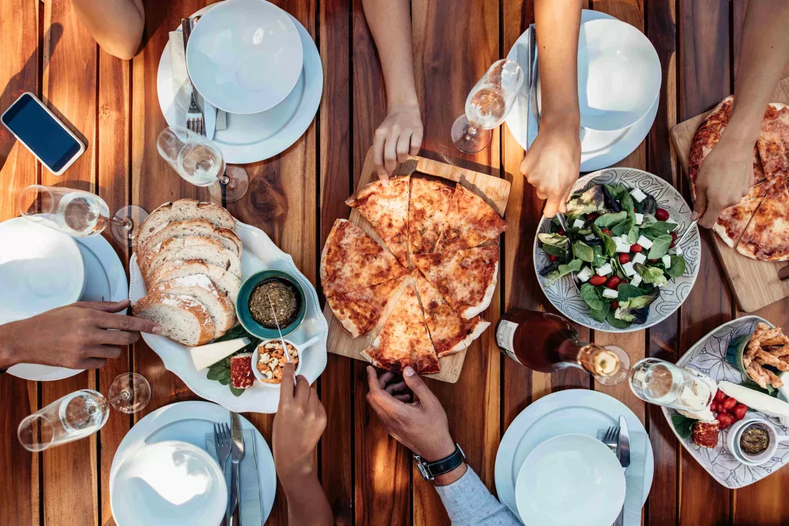A top-down view of a wooden table set with pizza, salad, bread, and appetizers, surrounded by people sharing the meal.