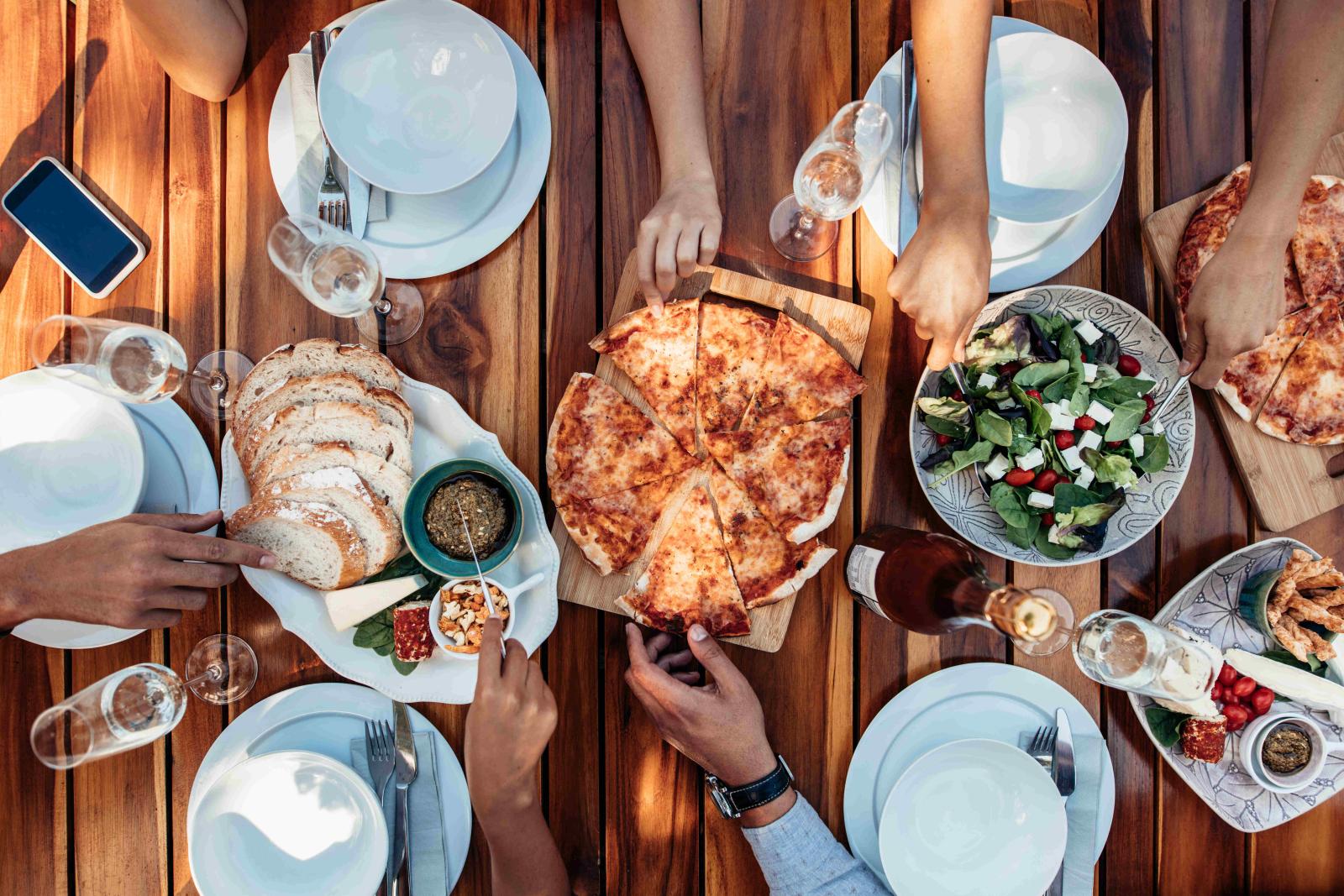 A top-down view of a wooden table set with pizza, salad, bread, and appetizers, surrounded by people sharing the meal.