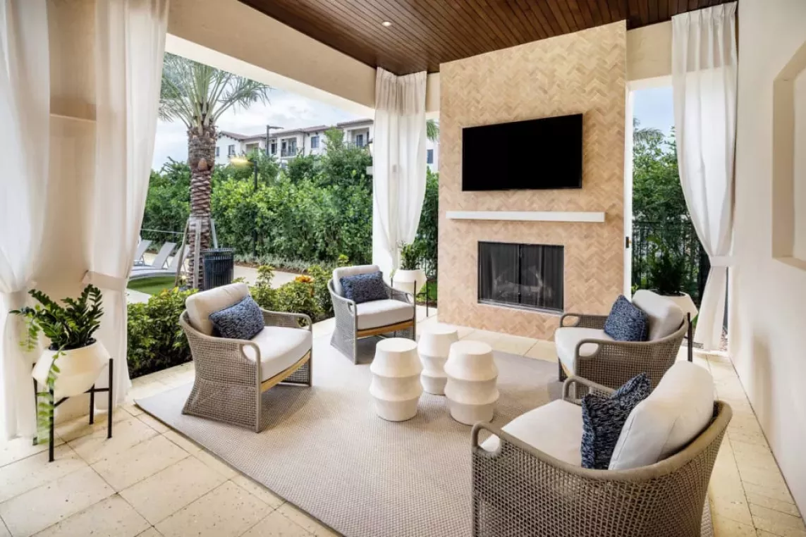 A luxurious covered seating area featuring a built-in fireplace, a mounted TV, and modern wicker chairs with blue cushions, surrounded by lush landscaping.