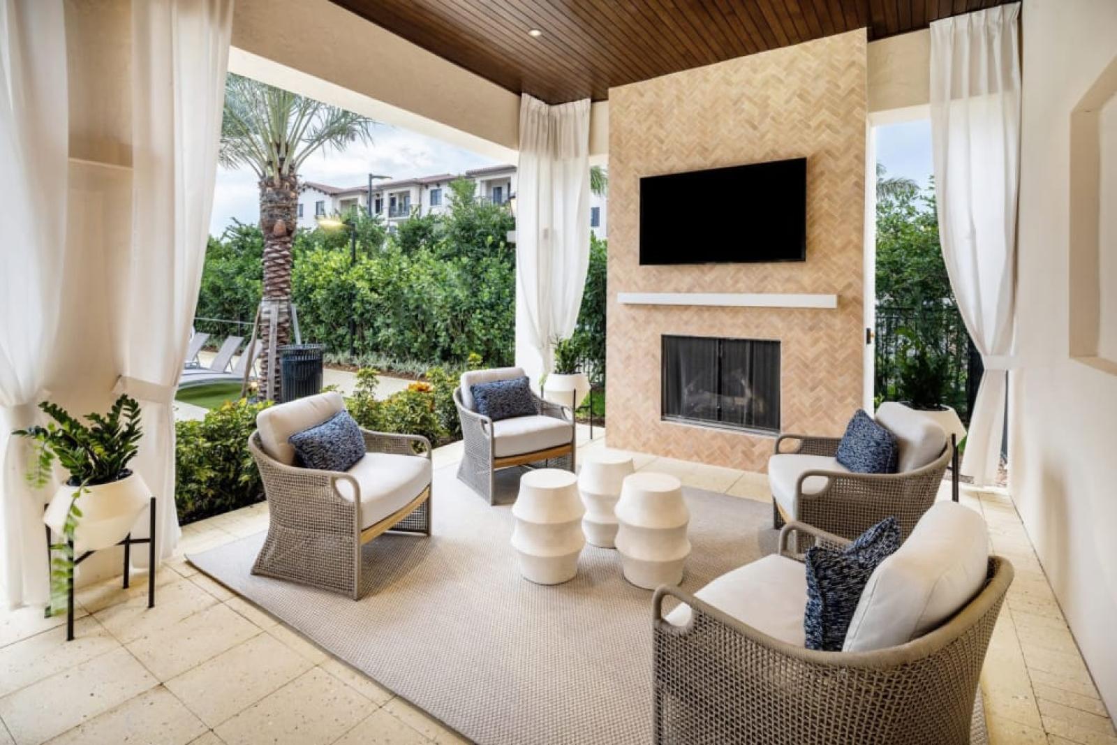 A luxurious covered seating area featuring a built-in fireplace, a mounted TV, and modern wicker chairs with blue cushions, surrounded by lush landscaping.