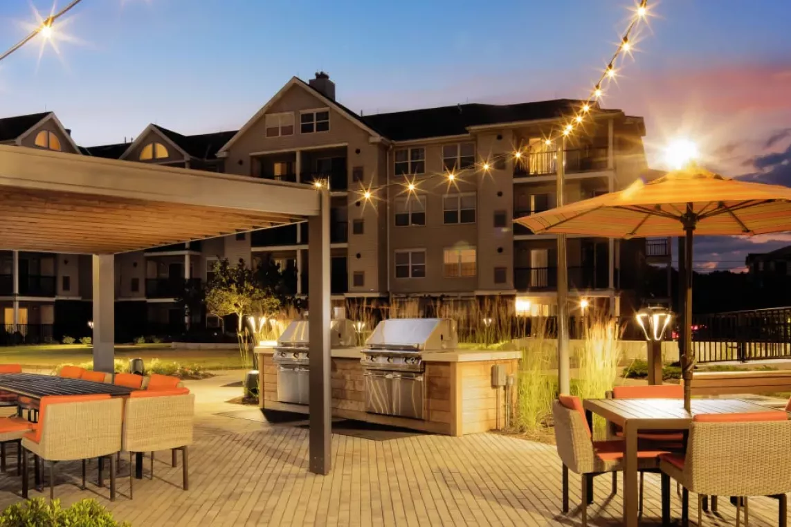A well-lit communal outdoor space with modern grilling stations, a covered dining area, and ambient string lighting for an inviting evening setting.