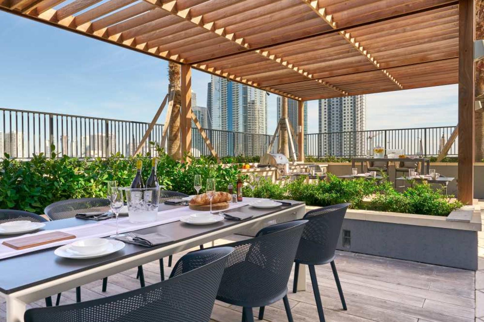 A rooftop dining area at Parkline Miami Luxury Apartments with a shaded wooden pergola, modern table settings, and lush greenery, offering skyline views.