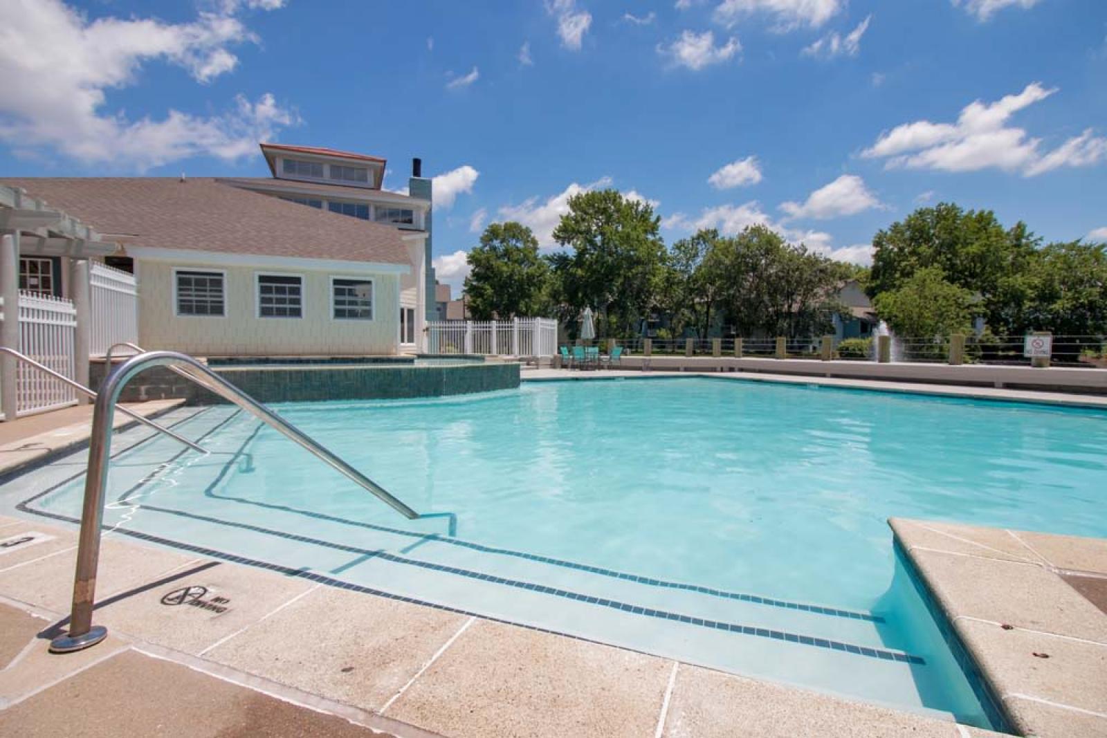 A sparkling outdoor pool with a stair entry, surrounded by a sunny deck, a poolside building, and lush trees under a bright blue sky.