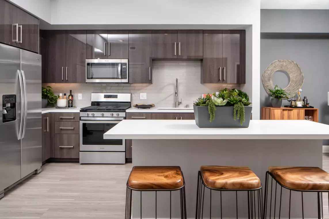 "Modern kitchen at The Pacific Luxury Apartments featuring sleek dark cabinetry, stainless steel appliances, a stylish white island with natural wood bar stools, and elegant décor accents."
