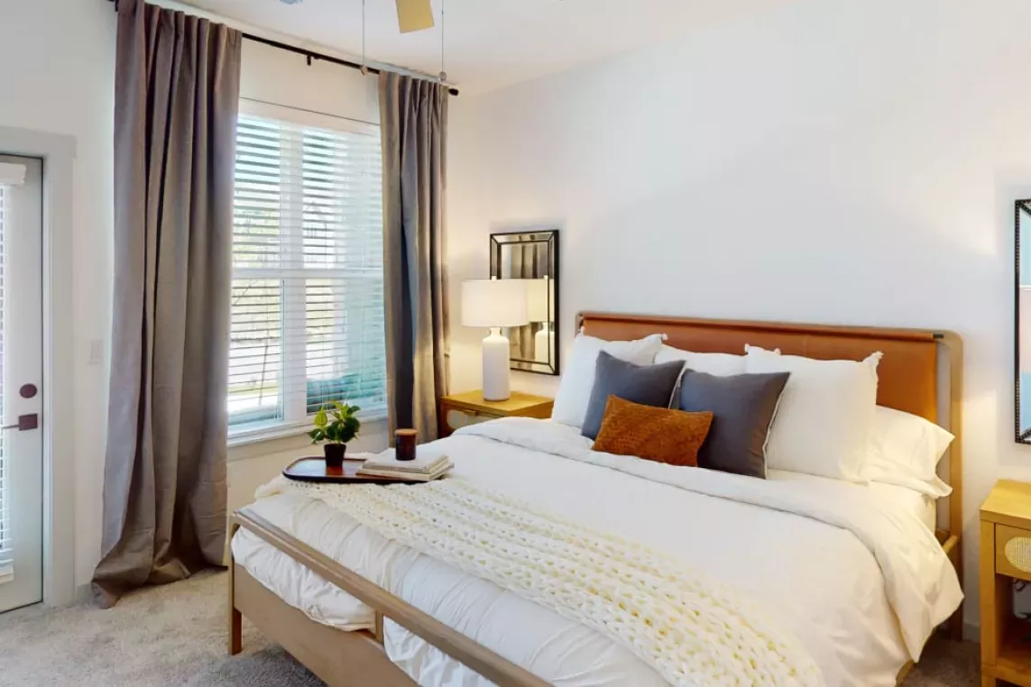 Spacious bedroom with a modern design, featuring a plush bed with layered pillows, bedside tables, and soft natural light through large windows.
