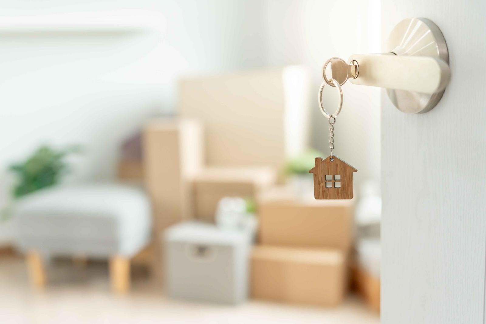 Close-up of a silver door handle with a key inserted, featuring a wooden house-shaped keychain. In the blurred background, a bright and modern living space is filled with moving boxes, a gray ottoman, and green plants, symbolizing a new home or apartment move-in.