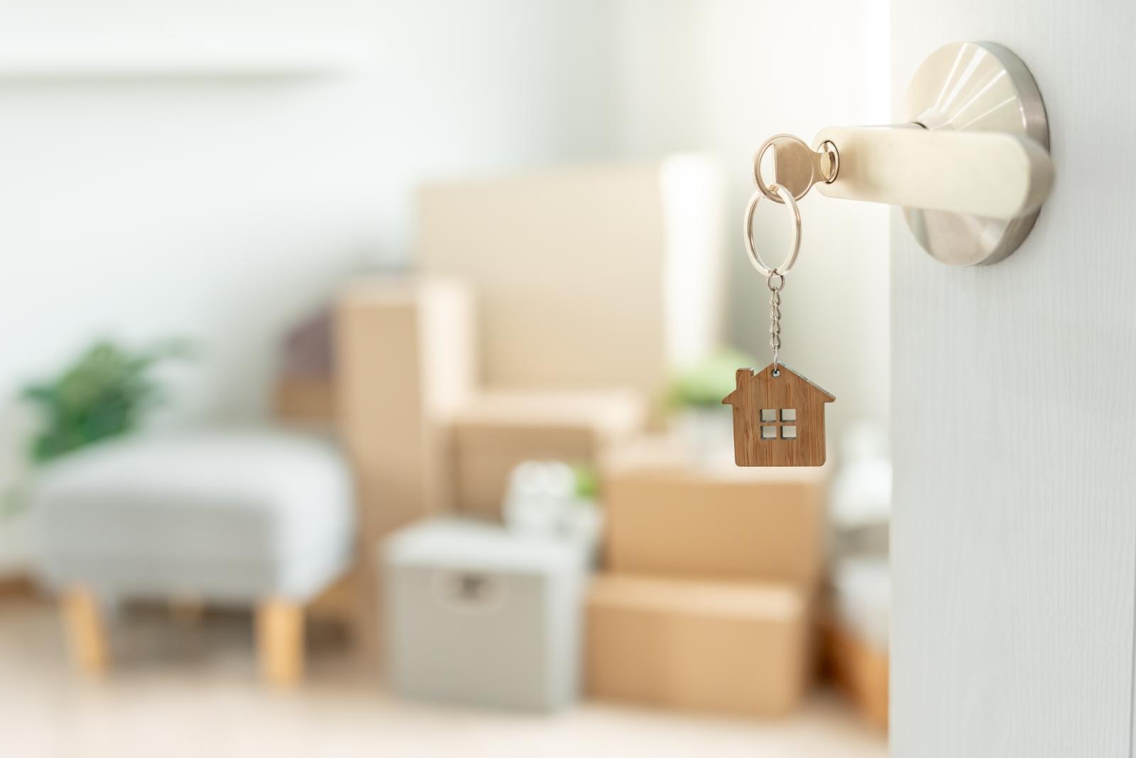 Close-up of a silver door handle with a key inserted, featuring a wooden house-shaped keychain. In the blurred background, a bright and modern living space is filled with moving boxes, a gray ottoman, and green plants, symbolizing a new home or apartment move-in.