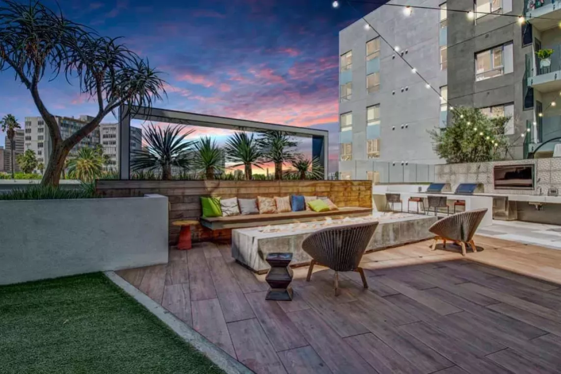 A sleek outdoor lounge area featuring a wooden bench with colorful cushions, a long fire pit, and ambient lighting against the cityscape.