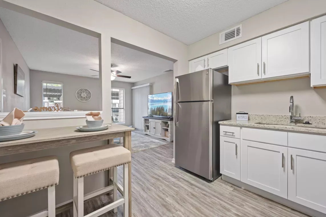 Bright, modern kitchen with white cabinetry, granite countertops, and stainless steel appliances, opening to a cozy dining nook with a wooden table and upholstered stools, and a living room with a ceiling fan and wall-mounted TV in view.