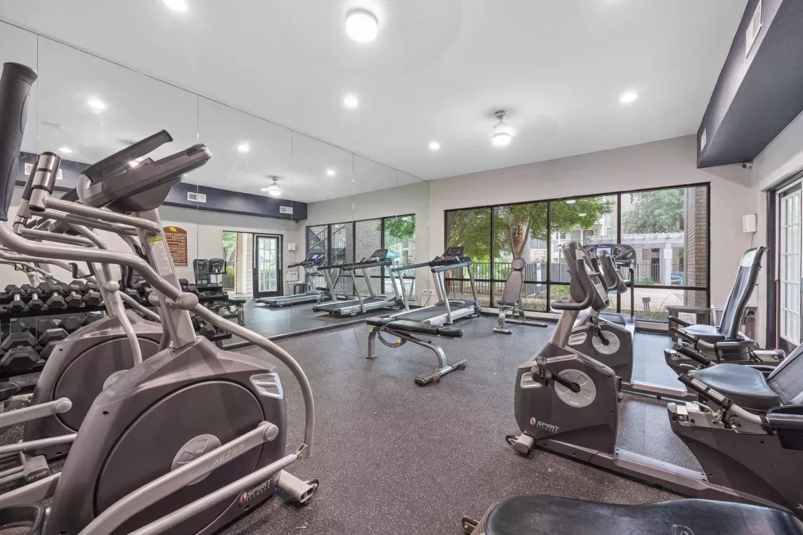 Spacious fitness center with mirrored walls, treadmills, ellipticals, recumbent bikes, free weights, and natural light from large windows overlooking the outdoor area.