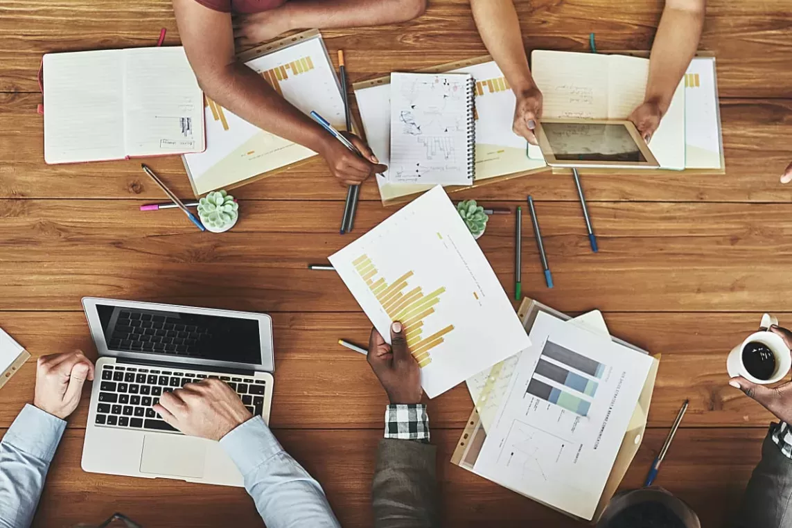 Top-down view of a diverse group of professionals collaborating around a wooden table in a modern office setting. They are analyzing data, discussing reports with charts and graphs, using a laptop and a tablet, and taking notes. The workspace is filled with pens, notebooks, and small potted plants, reflecting a dynamic and productive teamwork environment.