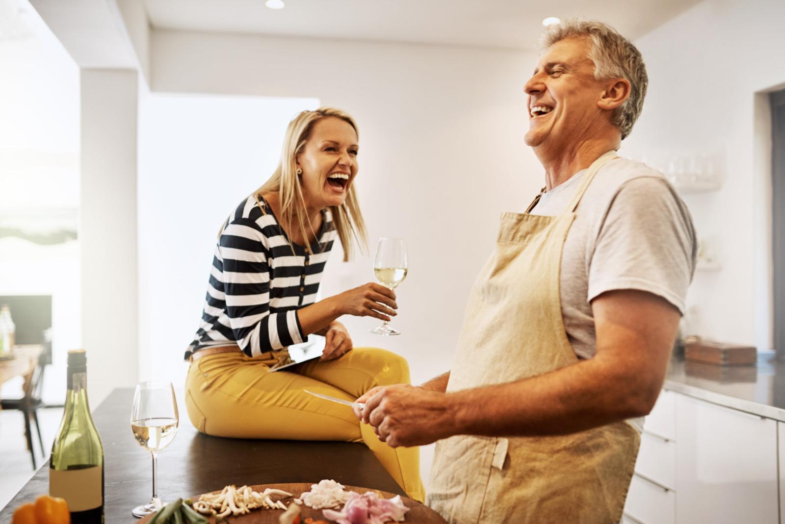 An older couple in the kitchen together laughing, drinking, and cooking together.
