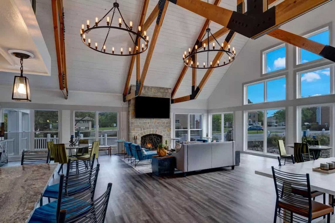 Modern clubhouse interior with vaulted ceilings, exposed wooden beams, and large windows letting in natural light. Features cozy seating around a stone fireplace, elegant chandeliers, and a mix of stylish tables and chairs, creating a warm and inviting communal space.