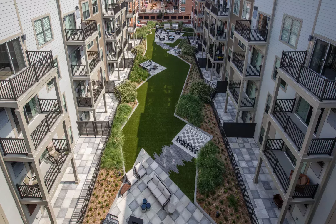 Aerial view of a modern apartment community courtyard featuring a central green space lined with outdoor lounge areas, a life-size chessboard, and firepit seating. The surrounding buildings have private balconies and neutral-toned siding, offering a sleek, contemporary aesthetic. The landscaping includes ornamental grasses and neatly maintained garden beds, creating an inviting atmosphere for residents to relax and socialize.