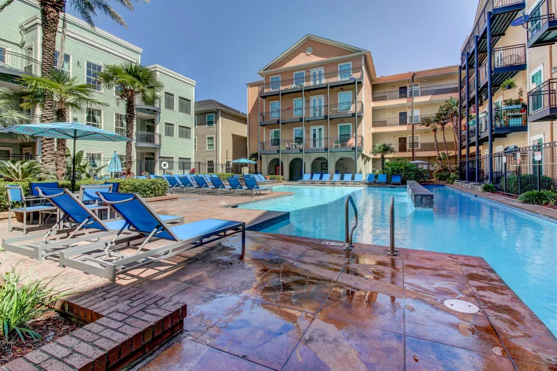 Luxury apartment community with resort-style pool, sun loungers, palm trees, and multi-story buildings featuring private balconies and shaded walkways.