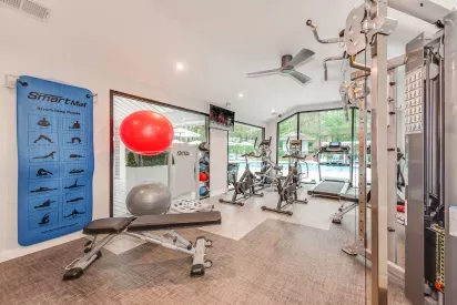 A bright fitness center equipped with exercise machines, stretching mats, stability balls, and large windows overlooking an outdoor pool area.
