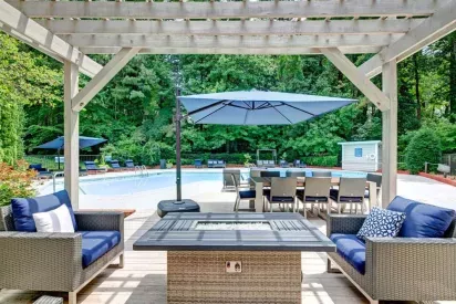 An inviting outdoor seating area with wicker furniture, a fire pit, and a view of the pool, surrounded by lush trees.