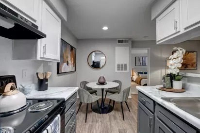 A modern kitchen with gray and white cabinetry, a marble-style countertop, and a dining nook featuring a round table with four upholstered chairs, accented by a circular mirror and abstract artwork on the walls.