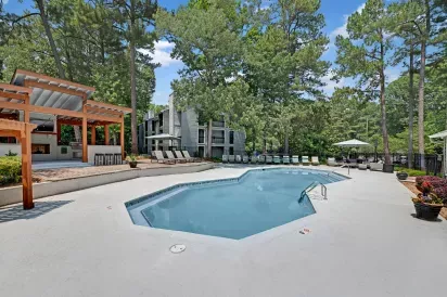 A spacious outdoor pool surrounded by lounge chairs, shaded seating areas, and tall trees, with an adjacent pergola providing additional seating.