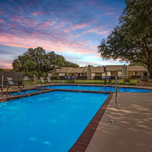 An outdoor pool illuminated at dusk, surrounded by lounge chairs, trees, and residential buildings under a vibrant sunset sky.