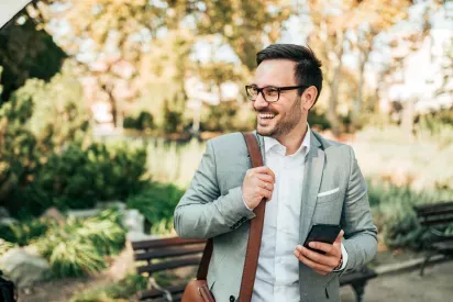  A professional man in a light gray blazer and glasses walking through a park, holding a smartphone and a leather bag. The scene highlights a modern, confident lifestyle surrounded by greenery and natural light.