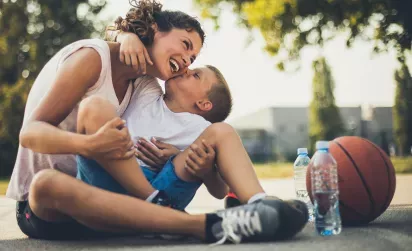 A mother and her young son share a joyful moment, laughing and hugging while sitting on a basketball court with a basketball and water bottles nearby.