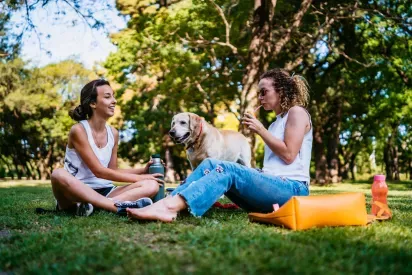  Two smiling women enjoying a picnic in a lush green park, accompanied by a friendly dog. The relaxed outdoor setting highlights a pet-friendly and community-oriented lifestyle under a bright, sunny day.