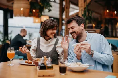  A smiling couple enjoying a meal together at a cozy restaurant, sharing food and laughter. The warm lighting and inviting atmosphere highlight a fun and relaxed dining experience.