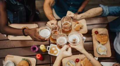 A group of people clinking beer mugs over a table with food, including burgers and onion rings.