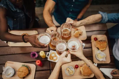 A group of friends raising beer mugs in a celebratory toast, seated at a wooden table with burgers and snacks.