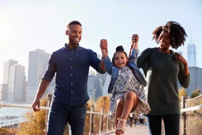 A joyful family holding hands, lifting their young daughter playfully, with a city skyline in the background.