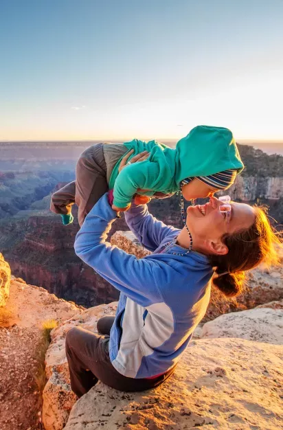 A woman playfully lifts a bundled-up child while sitting on a rocky ledge, with a breathtaking canyon landscape and sunset in the background.